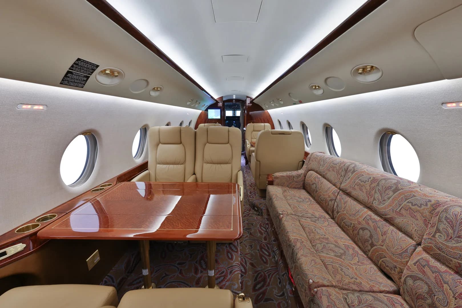 The spacious interior of a private jet features beige leather seats, a wooden table, and a patterned sofa, with large oval windows along the cabin.