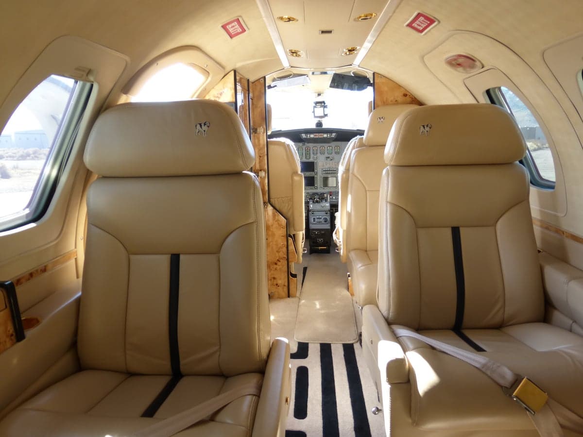 The interior cabin of a private jet, featuring tan leather seats and wood-paneled walls, looking towards the cockpit.