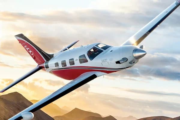 A white and red Piper M600 single-engine turboprop aircraft flying against a sunset sky over mountains.
