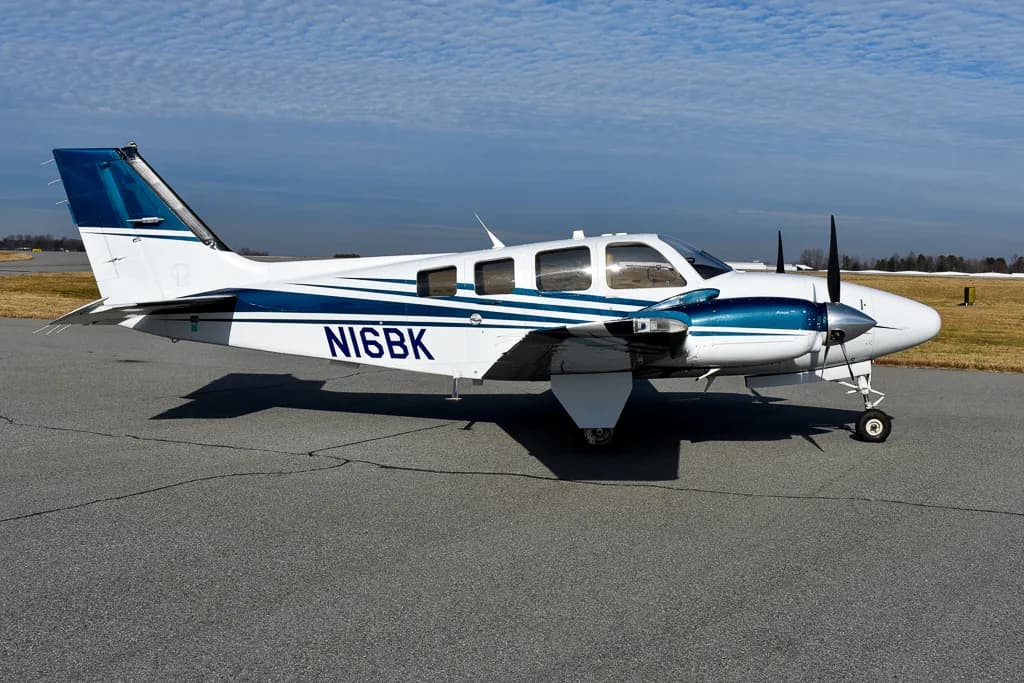 A white and dark blue Beechcraft Bonanza, N16BK, is parked on an asphalt runway with a grassy field in the background.