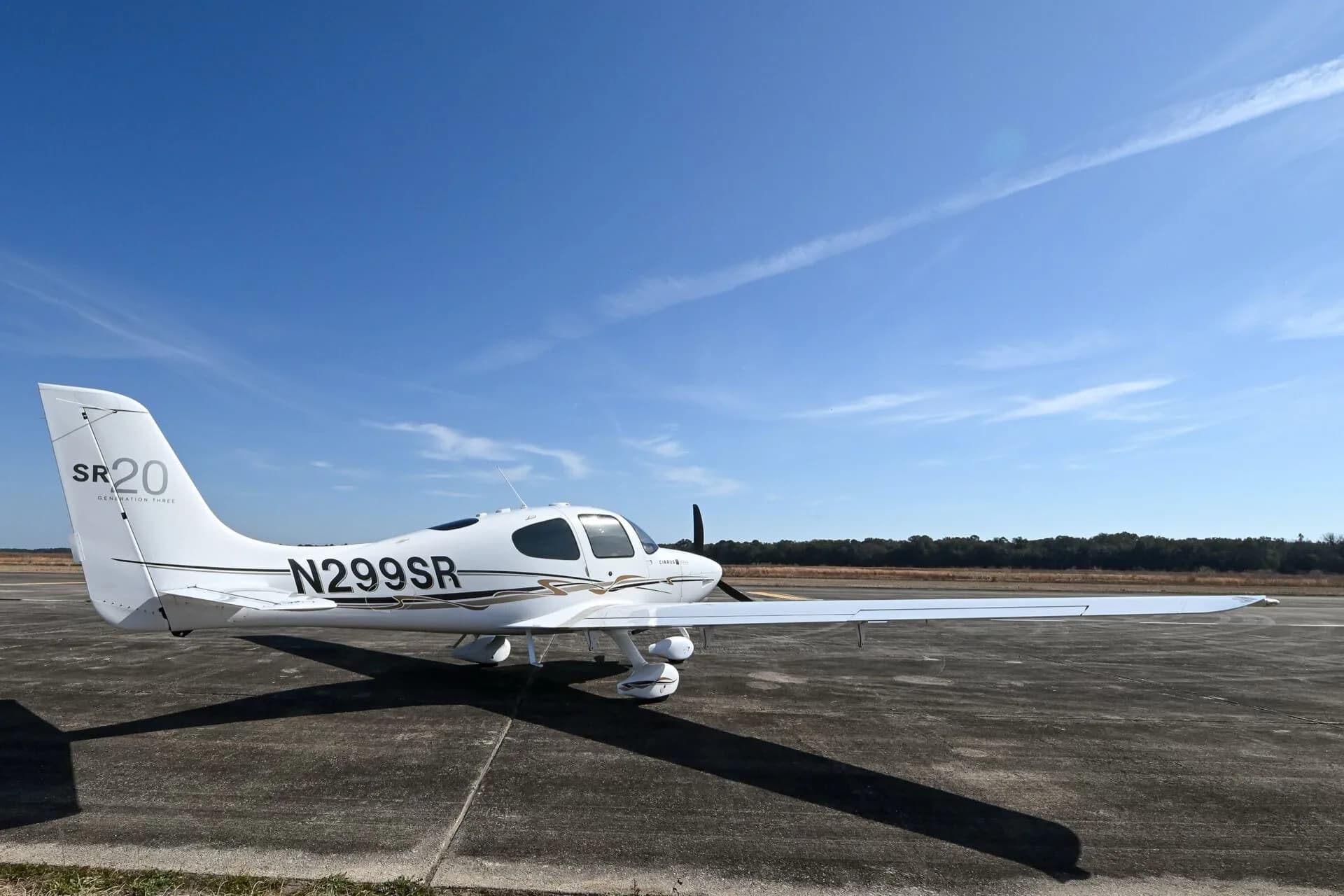 A white Cirrus SR20 aircraft with black and gold accent stripes, registration N299SR, parked on an airfield under a clear blue sky.