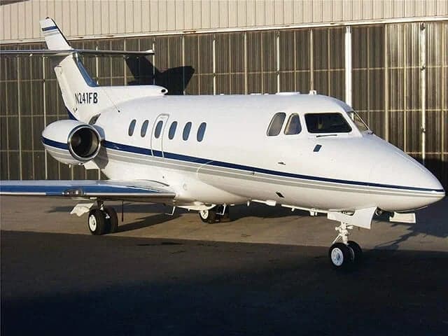 A white business jet with blue and grey stripes parked on an airport tarmac in front of a hangar.