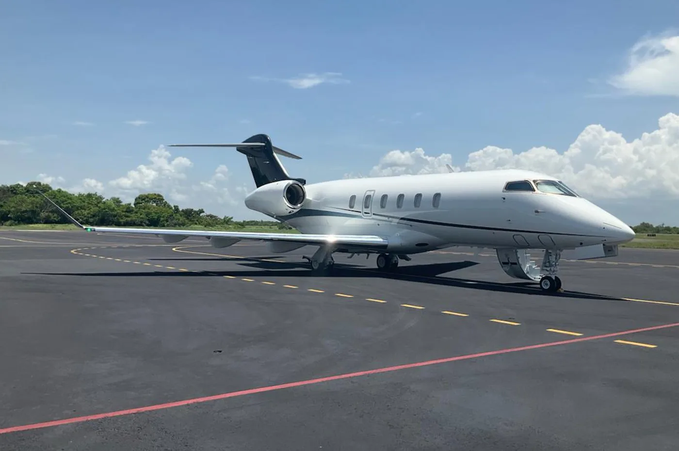 A white Bombardier Challenger business jet with black tail accents is parked on an asphalt tarmac under a blue sky with scattered clouds.