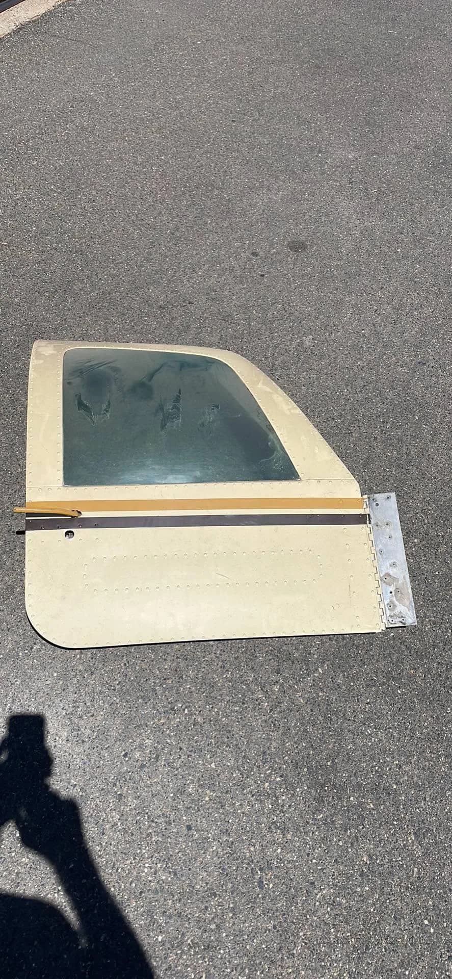 A detached aircraft panel with a small window and hinge, lying on asphalt.