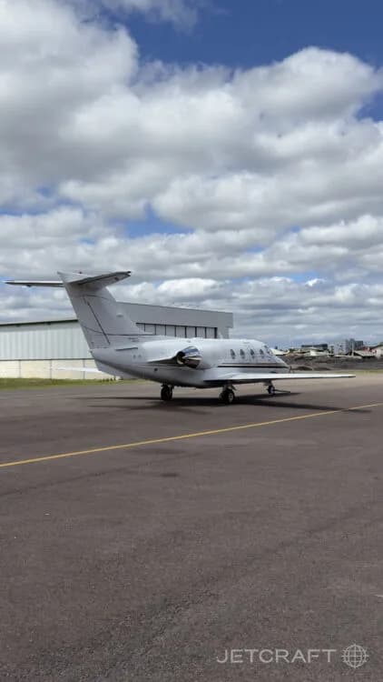 A white Hawker 800XP business jet parked on an airport tarmac with a hangar in the background.