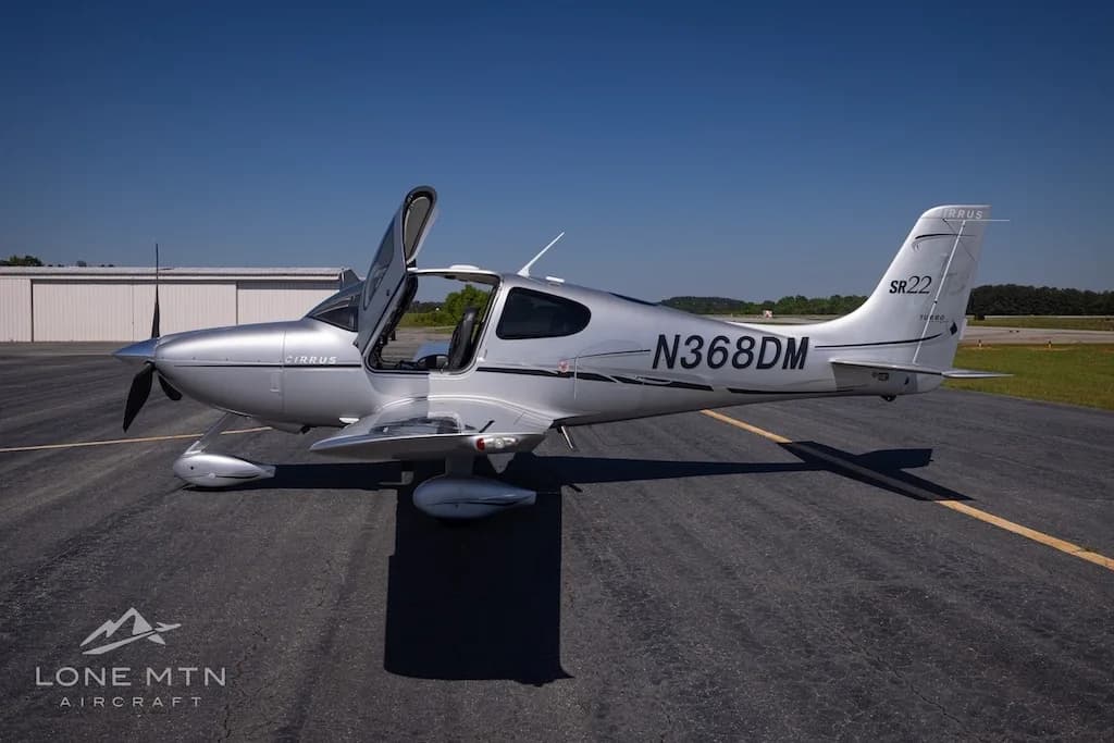 A silver Cirrus SR22 Turbo aircraft (N368DM) with its doors open, parked on an asphalt runway under a clear blue sky.