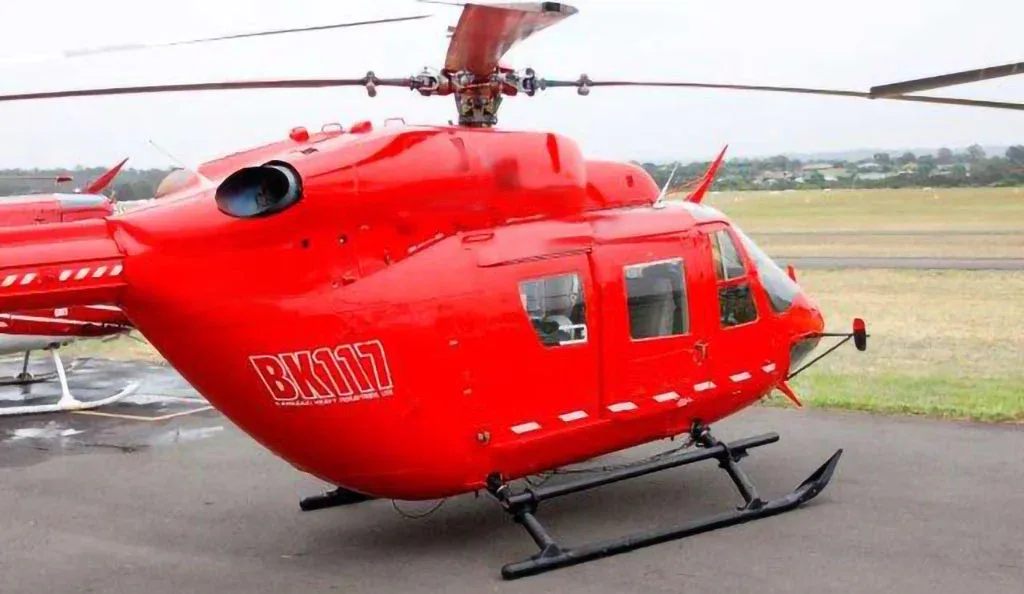 A red BK117 helicopter with 'BK117' on its side, parked on an airfield.