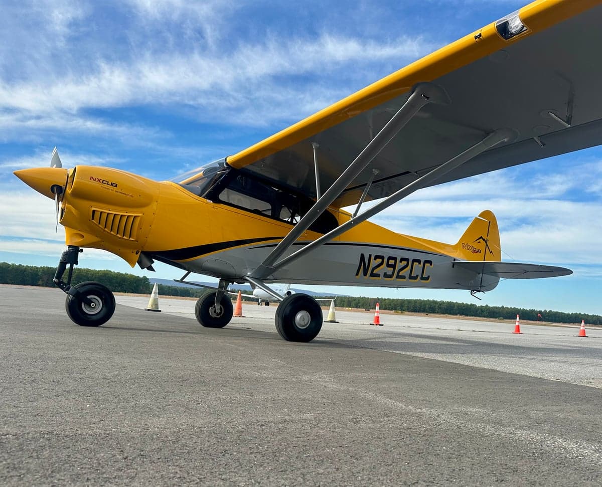 A yellow and gray NX Cub aircraft, N292CC, parked on an asphalt tarmac under a blue sky.