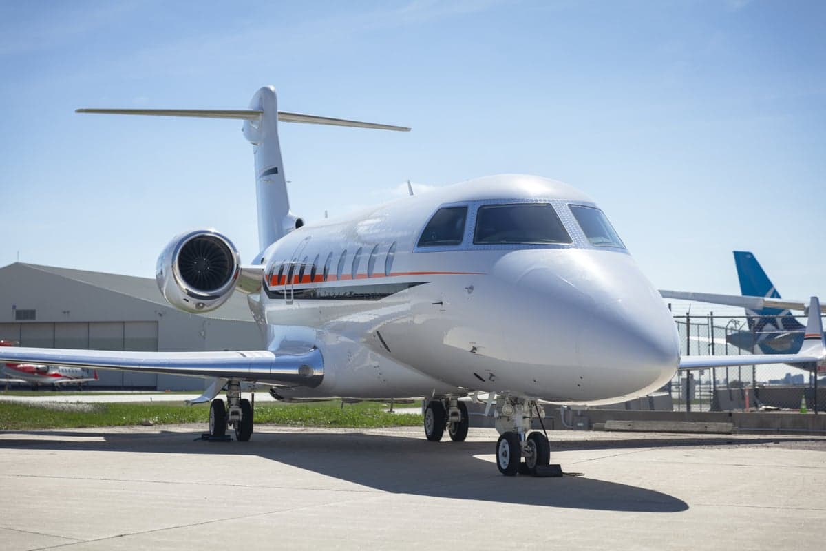 A white private jet is parked on the tarmac with a hangar in the background.