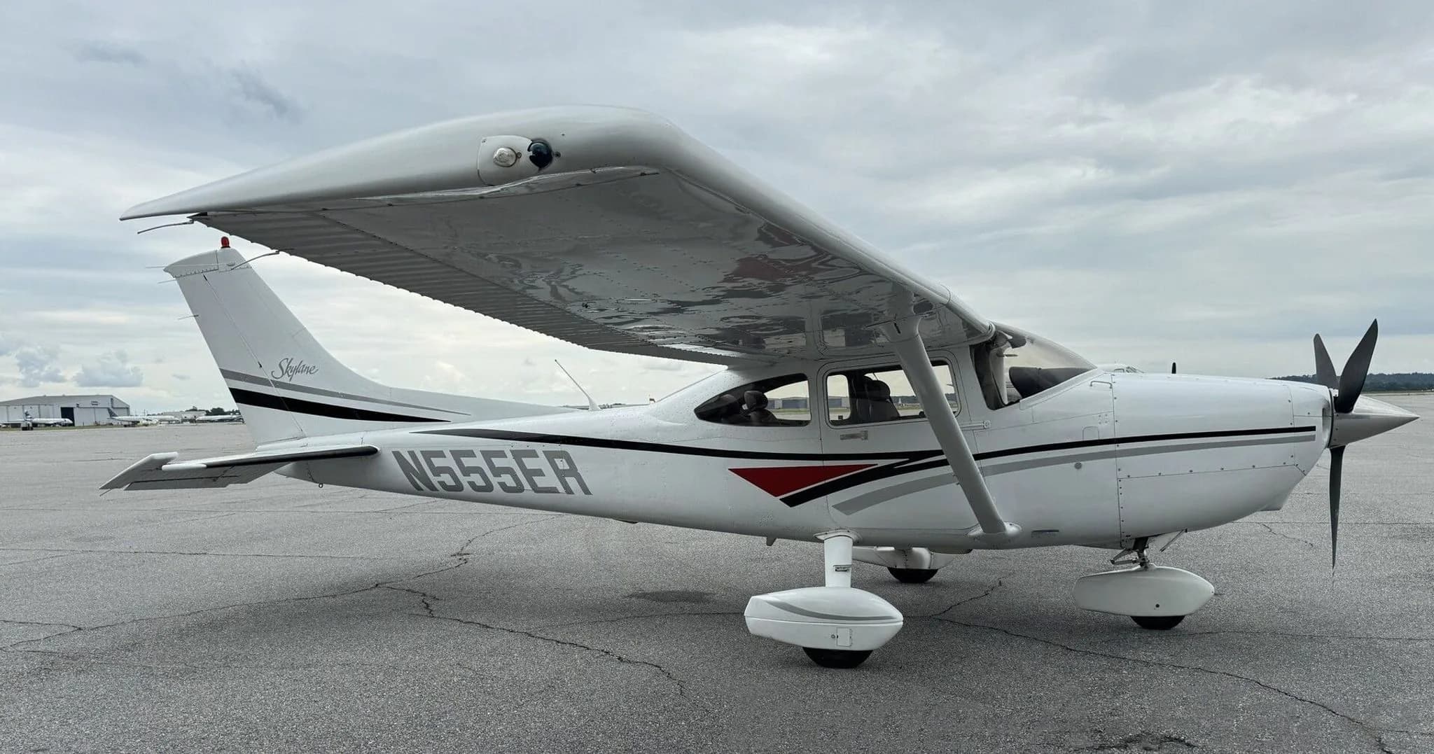 A white Cessna 182 Skylane with black, grey, and red stripes parked on an airfield under a cloudy sky.
