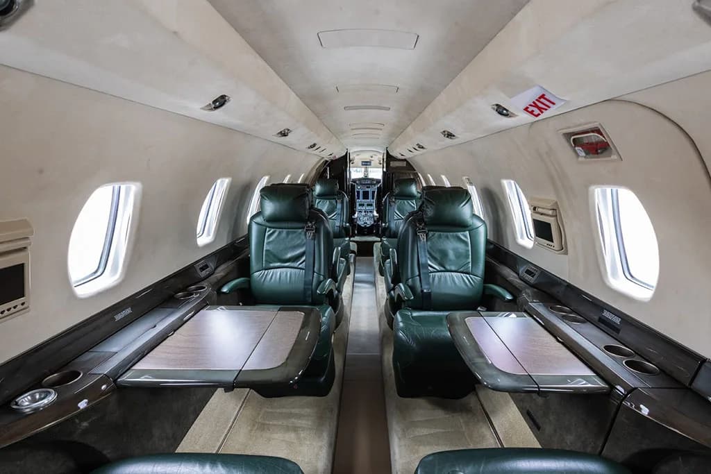 The interior cabin of a private jet with dark green leather seats and tables deployed, looking towards the cockpit.