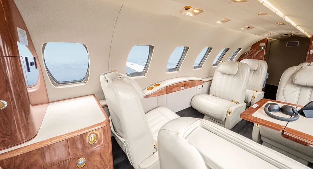 A side view of the aircraft cabin interior with light cream leather seats, wooden cabinetry, and multiple windows.