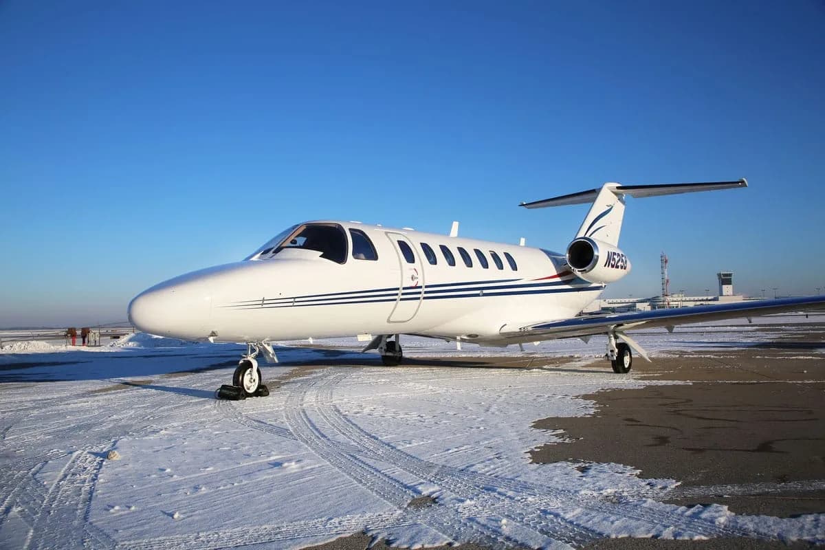 A white Cessna CitationJet with blue stripes, registration N525B, parked on a snowy tarmac under a clear blue sky, viewed from the front-right.