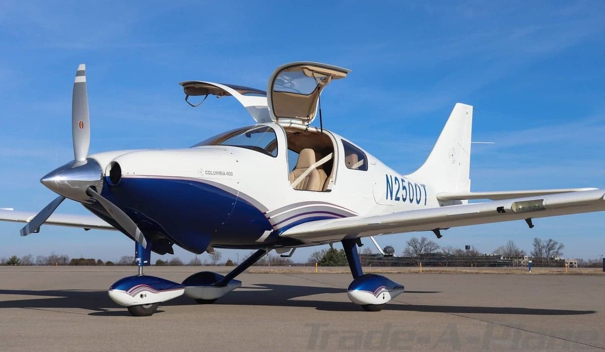 Three-quarter front view of a white and blue Columbia 400 aircraft with its gull-wing doors open on an airfield.