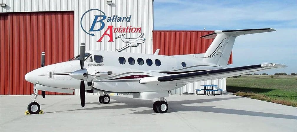 Side view of a Beechcraft King Air B200 parked in front of a hangar with the Ballard Aviation logo.