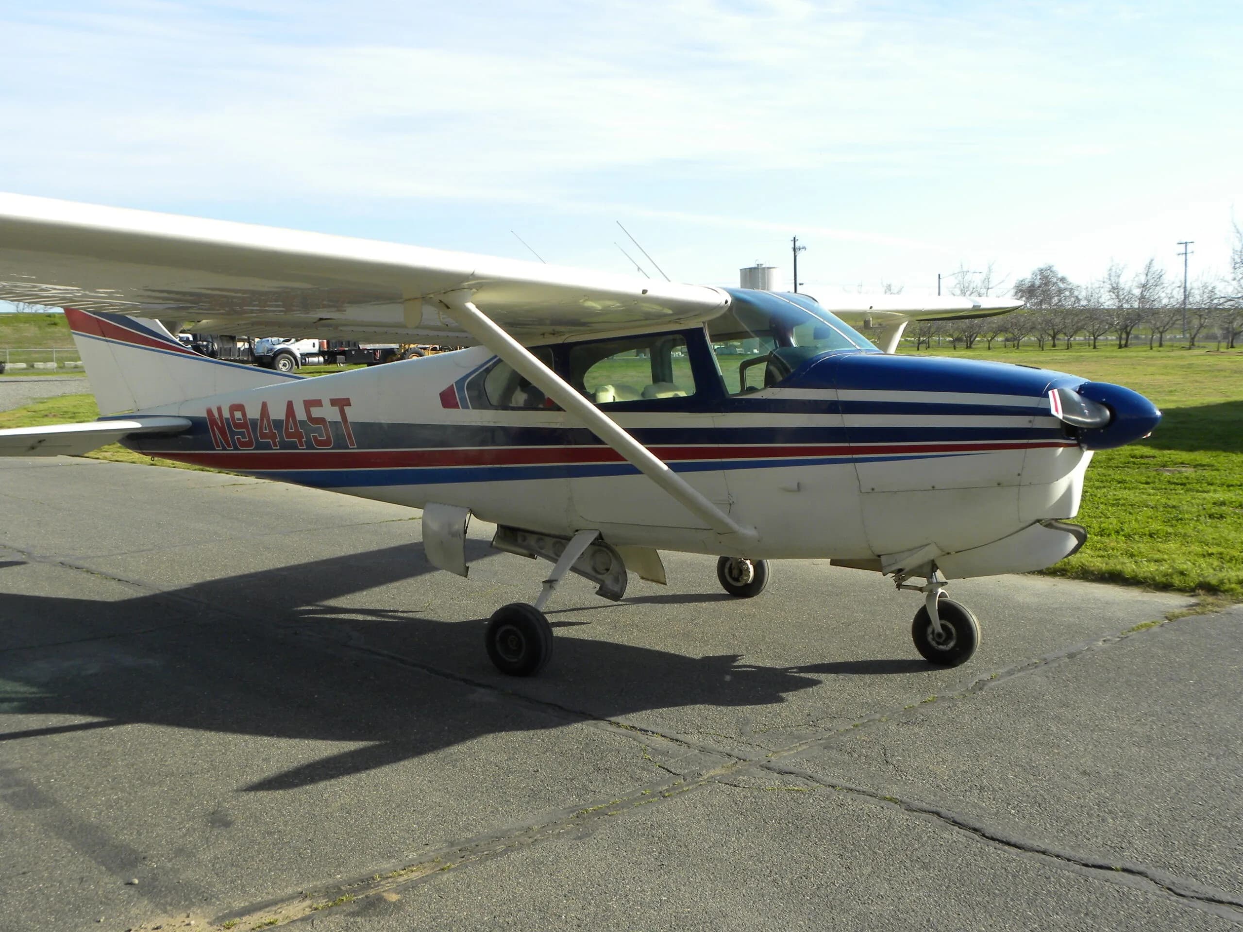 A side view of a white Cessna 182 with blue and red stripes, registration N9445T, on a tarmac.