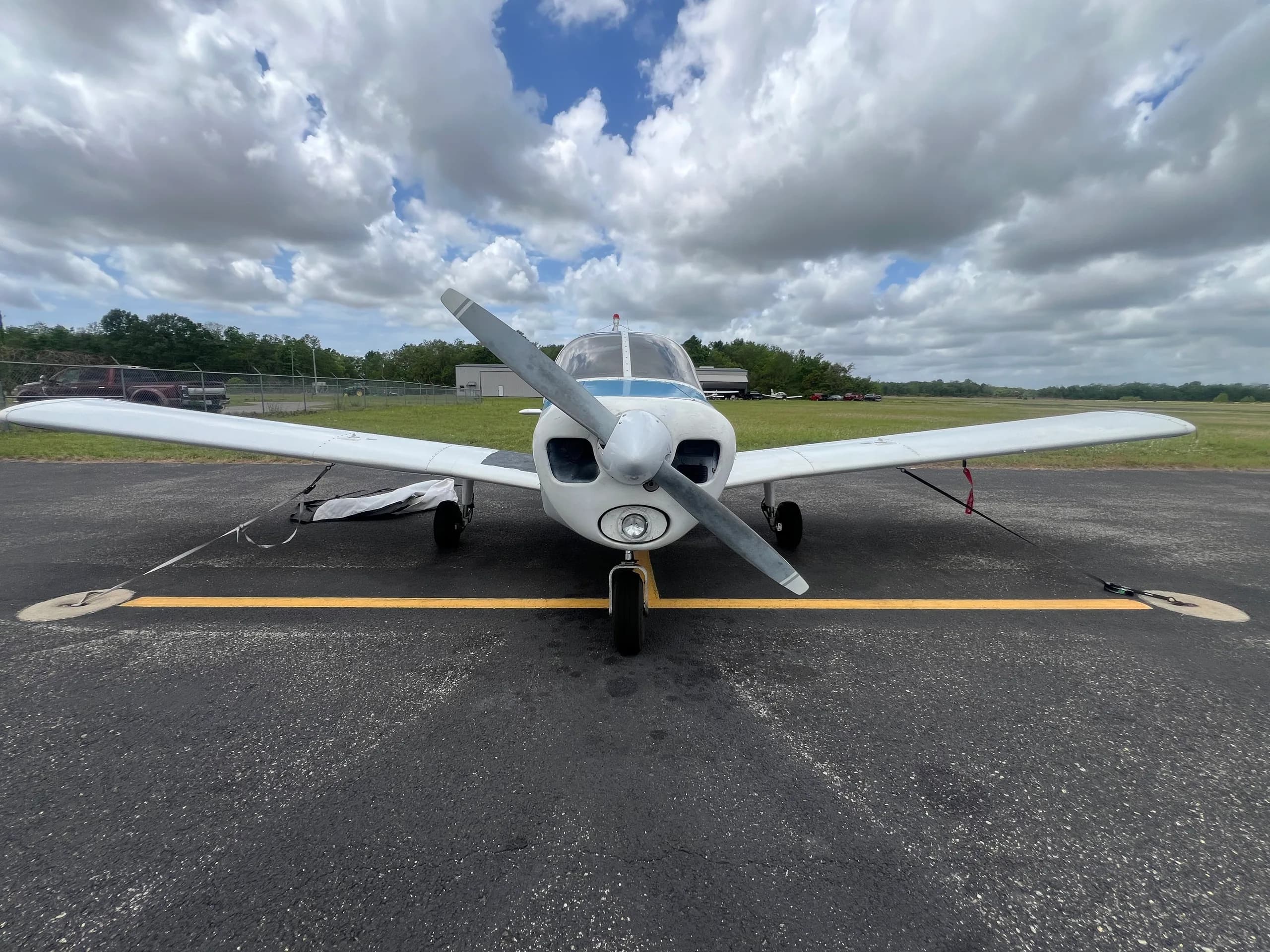 Front view of a white and blue Piper Cherokee 140 parked on an asphalt ramp under a cloudy sky.