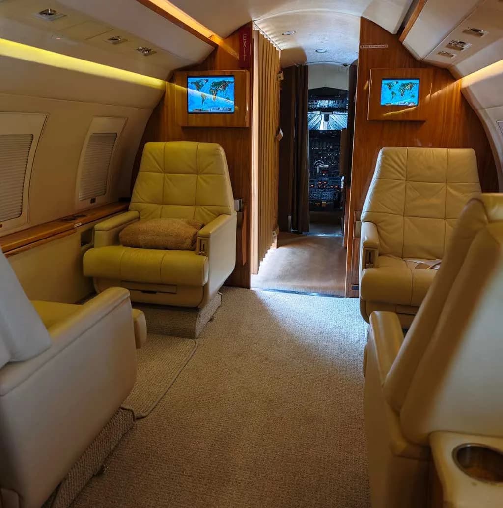 The business jet's cabin interior with beige leather seats, wood accents, and two screens displaying maps.