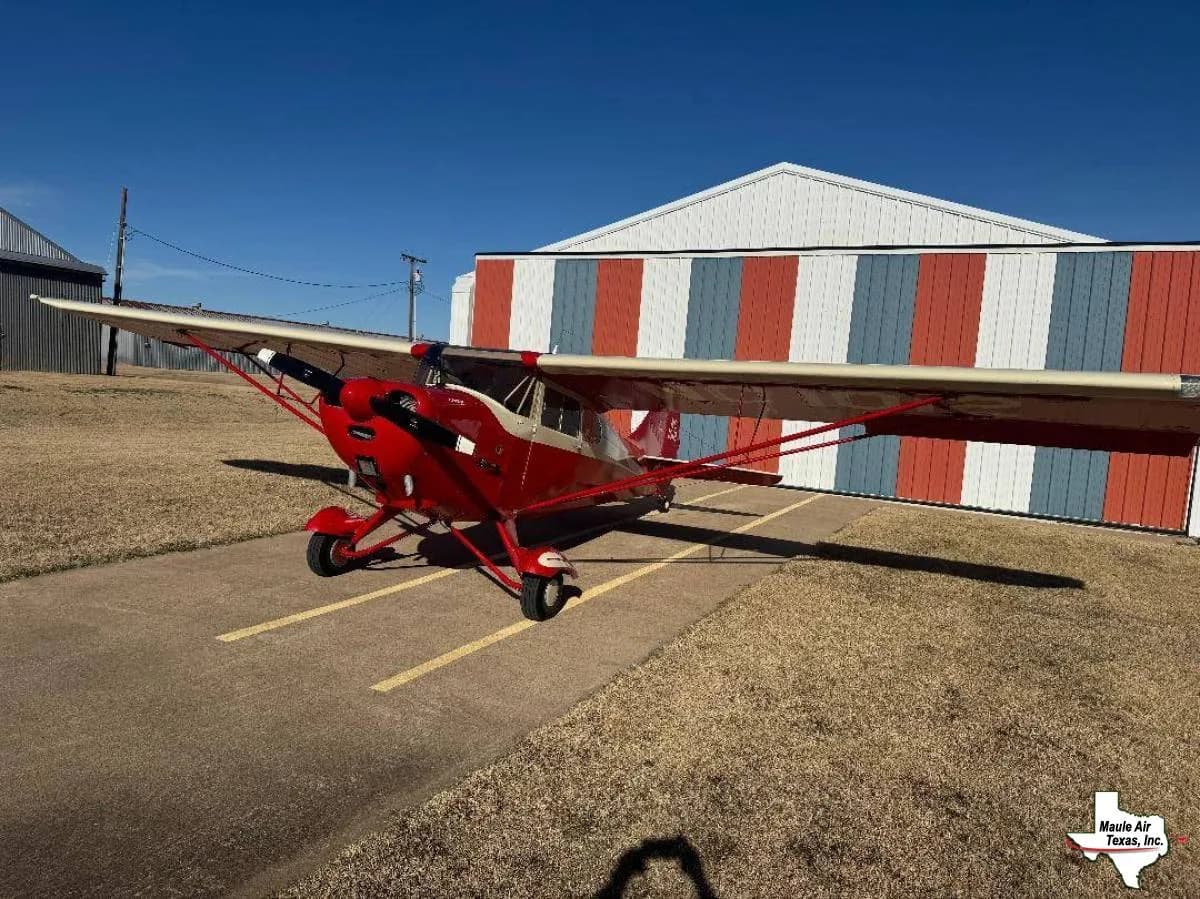 A red and cream high-wing taildragger aircraft parked on an airfield next to a striped hangar.