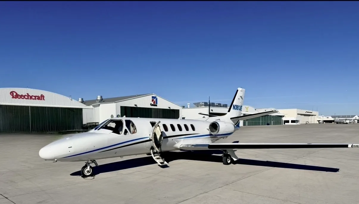 Front-left view of a white Cessna CitationJet with blue and black stripes, registration N361JE, parked on an airfield with its main door open, under a clear blue sky.