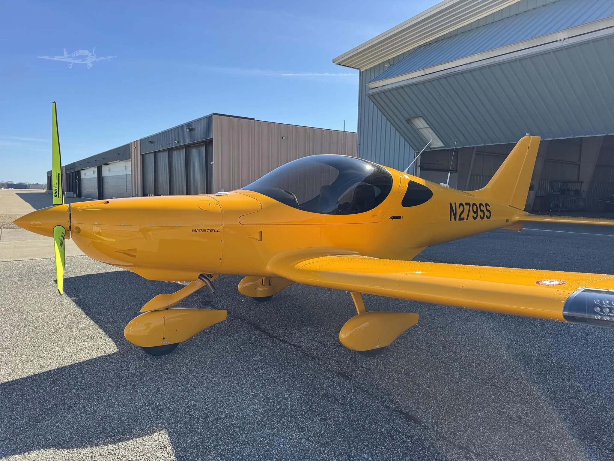 A bright yellow Bristell light sport aircraft, registration N279SS, parked on an asphalt ramp in front of hangars under a clear blue sky.