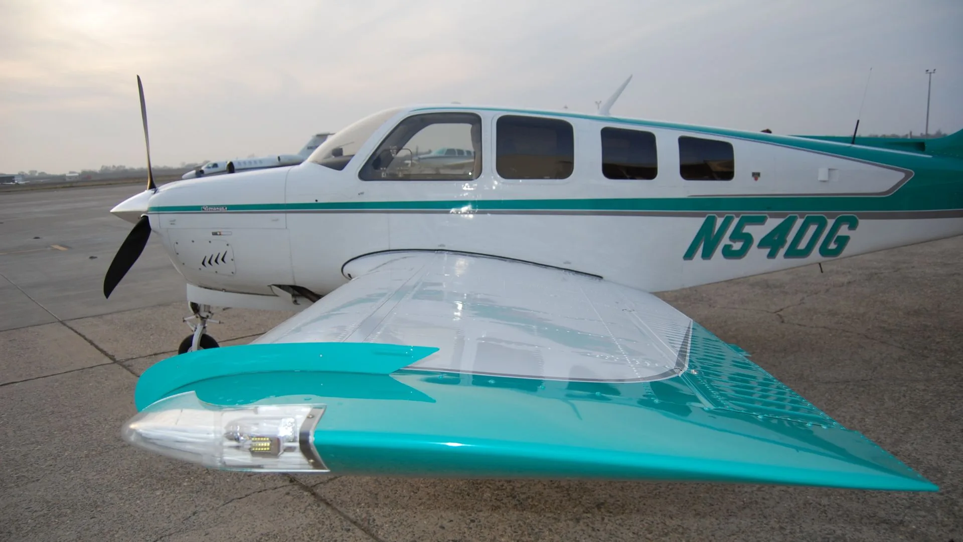 A close-up of the wing and fuselage of the Beechcraft Bonanza N54DG, showing the teal and white paint scheme and the registration number.