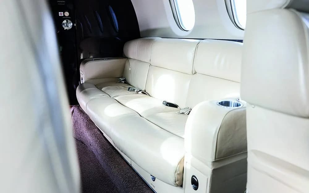 A cream-colored leather three-seat sofa with seatbelts and a cup holder in the aircraft cabin.