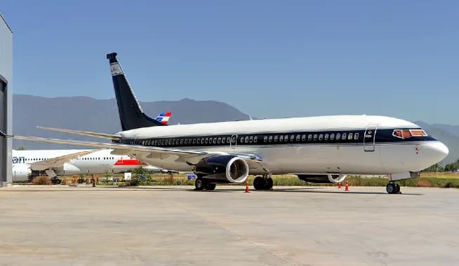 A Boeing 737 BBJ in a dark blue and white livery parked on an airfield with mountains in the background.