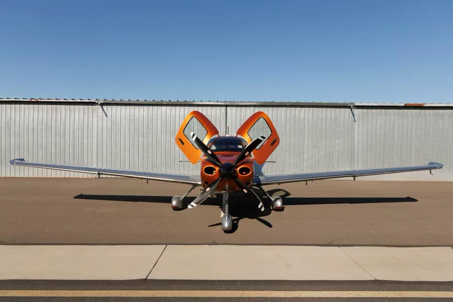 Front view of an orange and silver Cirrus SR22T GTS aircraft with both doors open, parked in front of a hangar.