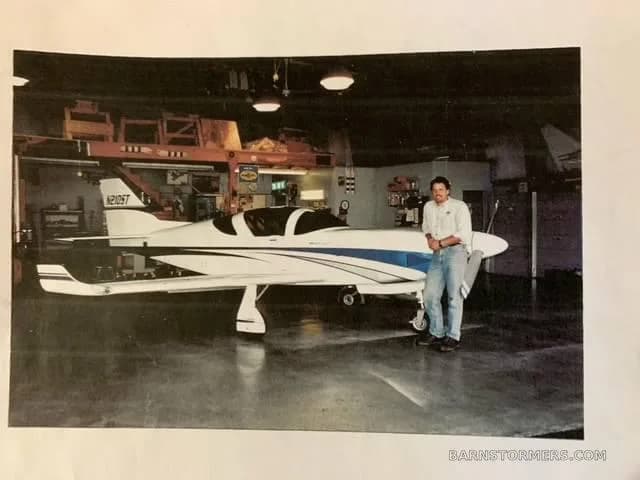 A man stands next to a white and blue single-engine aircraft (N210ST) inside a hangar, captured from a printed photo.