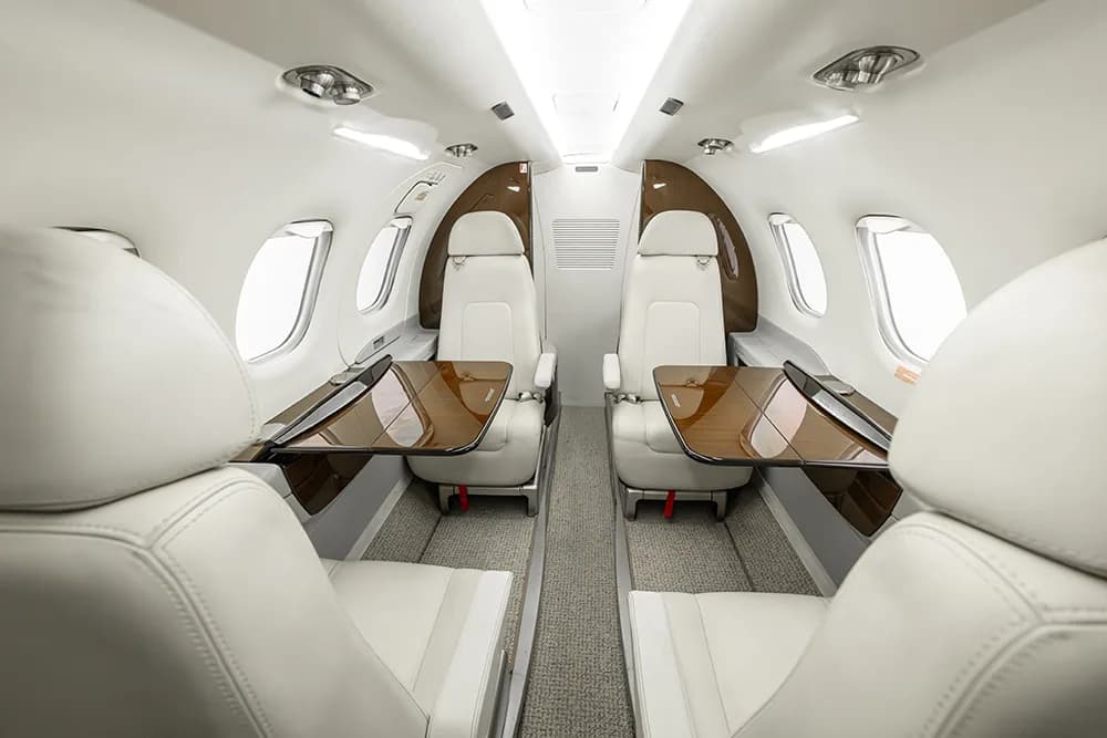 The interior of a private jet cabin showing white leather seats with dark wood tables deployed.