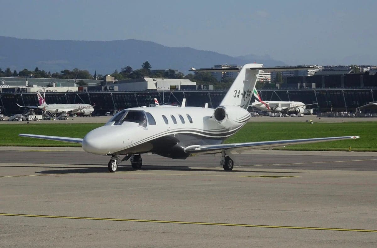 A white Cessna CitationJet with grey and black stripes, registration 3A-MSR, on a taxiway at a busy airport.