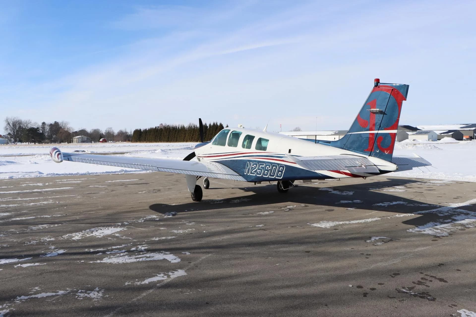 Side view of a white, blue, and red single-engine aircraft with registration N256DB on the fuselage, parked on a snowy tarmac.