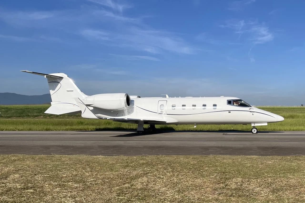 A white business jet with blue pinstripes is parked on a runway under a clear blue sky, viewed from the side.