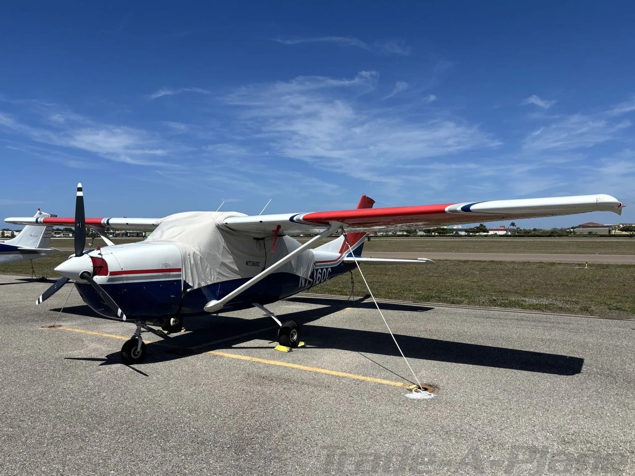 A white, blue, and red Cessna N7360C parked on an airfield with engine and cockpit covers, showing the N-number.