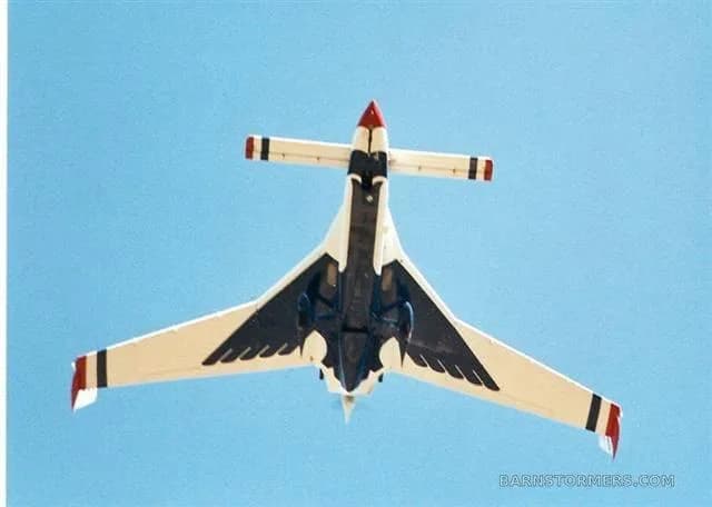 An unusual white, red, and blue aircraft with a delta-like wing and twin tails, viewed from directly below against a blue sky.