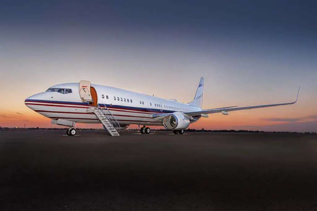 A white Boeing 737 with red and blue stripes, front door open with stairs, on the tarmac at sunset.
