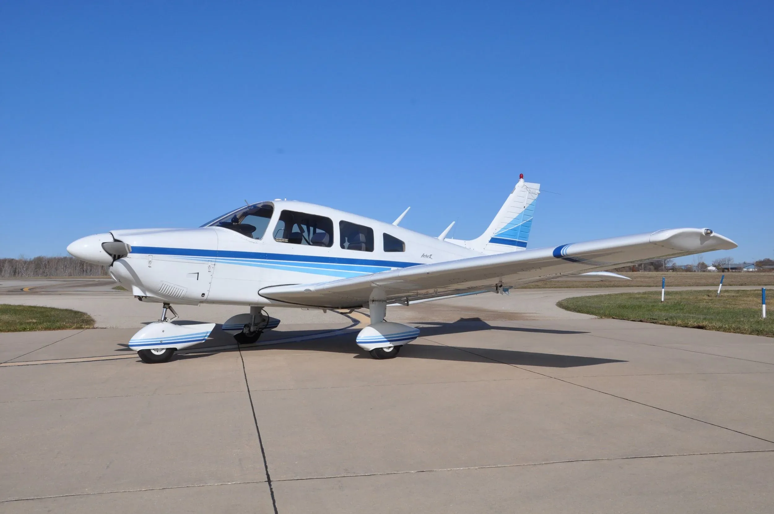 A white single-engine propeller aircraft with blue stripes parked on an asphalt surface under a clear blue sky.
