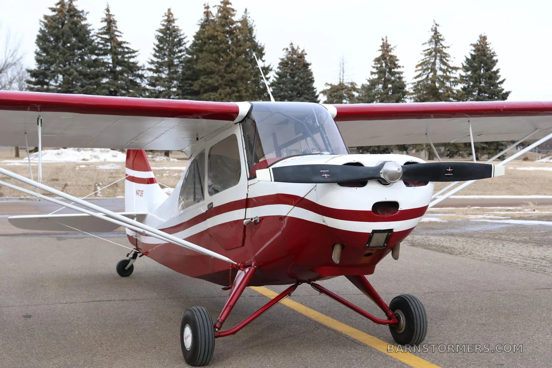 A front-right view of the red and white N4715E taildragger aircraft, showing its propeller and landing gear on an asphalt surface.