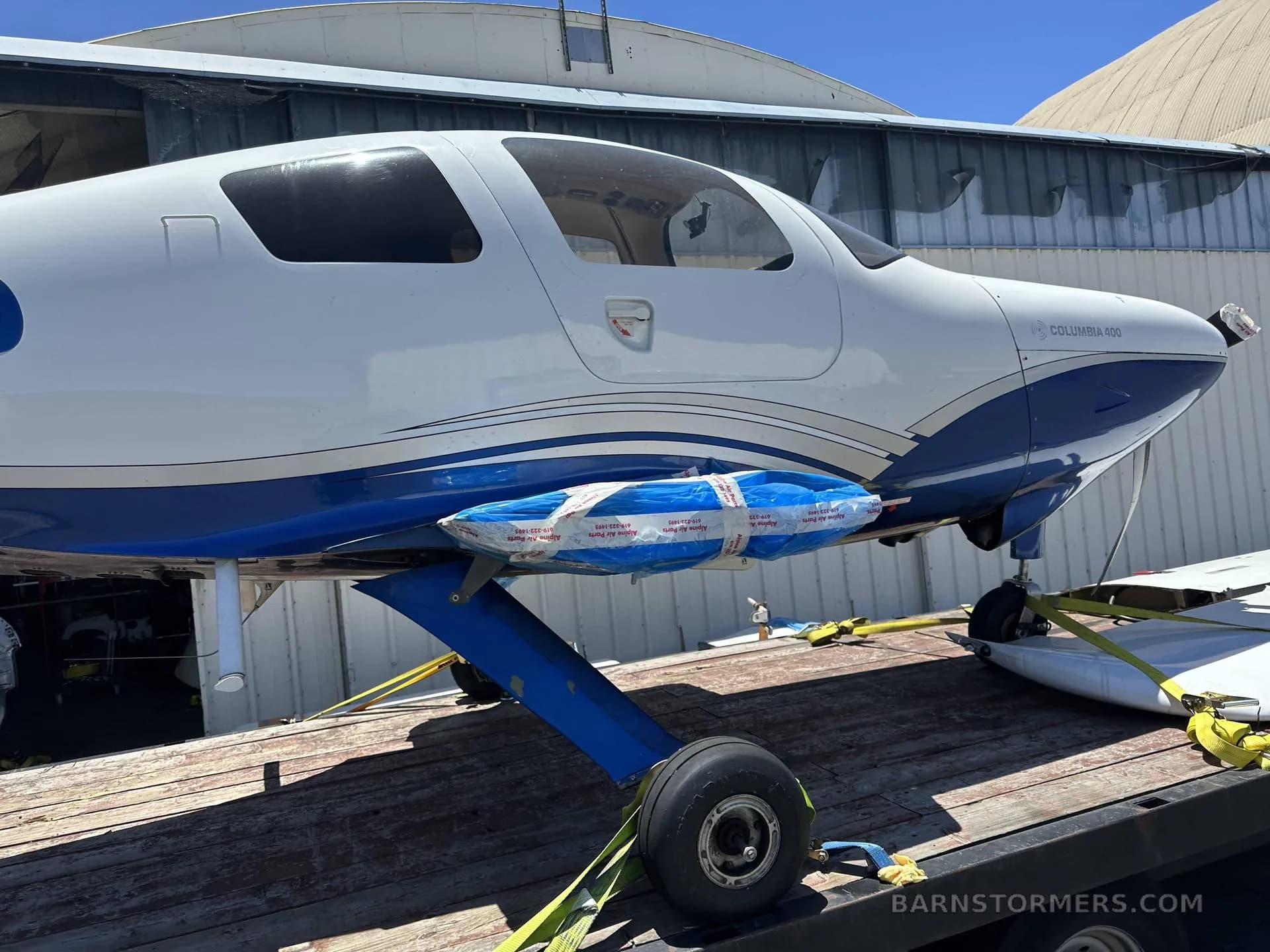 A side view of a white and blue Columbia 400 aircraft, showing the main landing gear and wrapped wing section, on a flatbed trailer.