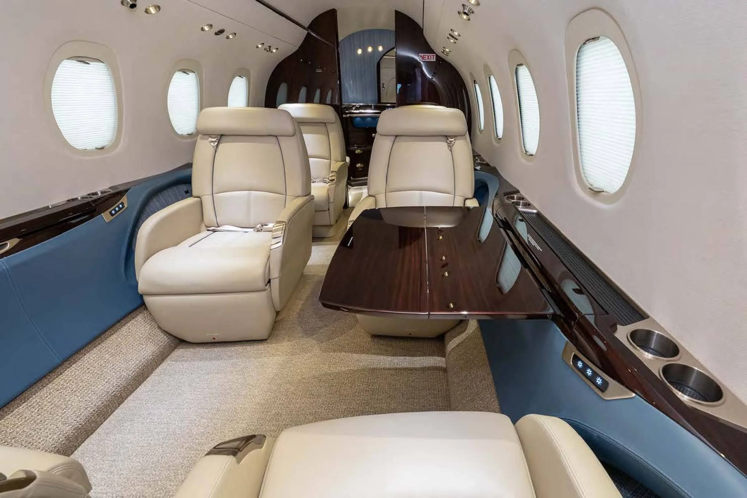 A view inside a private jet cabin showing beige leather seats, a polished wooden table, and blue side panels.