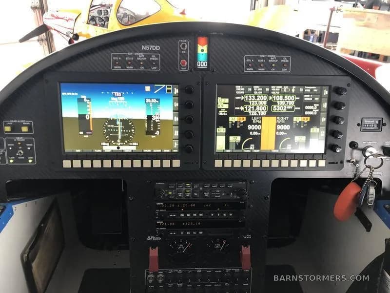 The cockpit instrument panel of a blue aircraft, featuring two large digital displays and various control switches.
