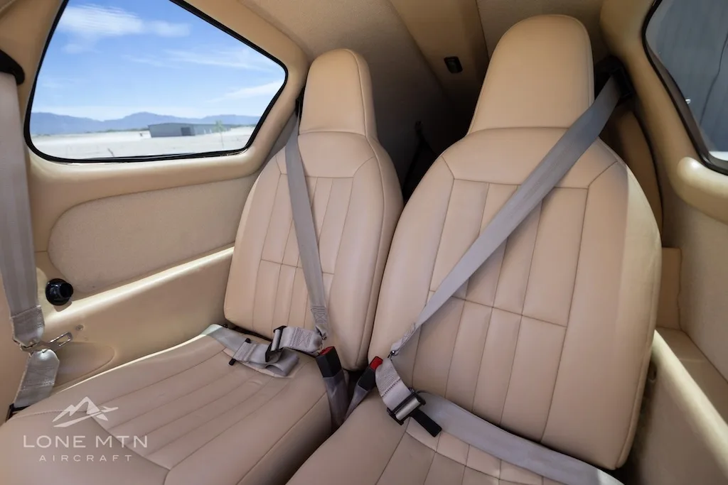 Two tan leather aircraft seats with seatbelts in the rear cabin, with a view of the outside through the window.