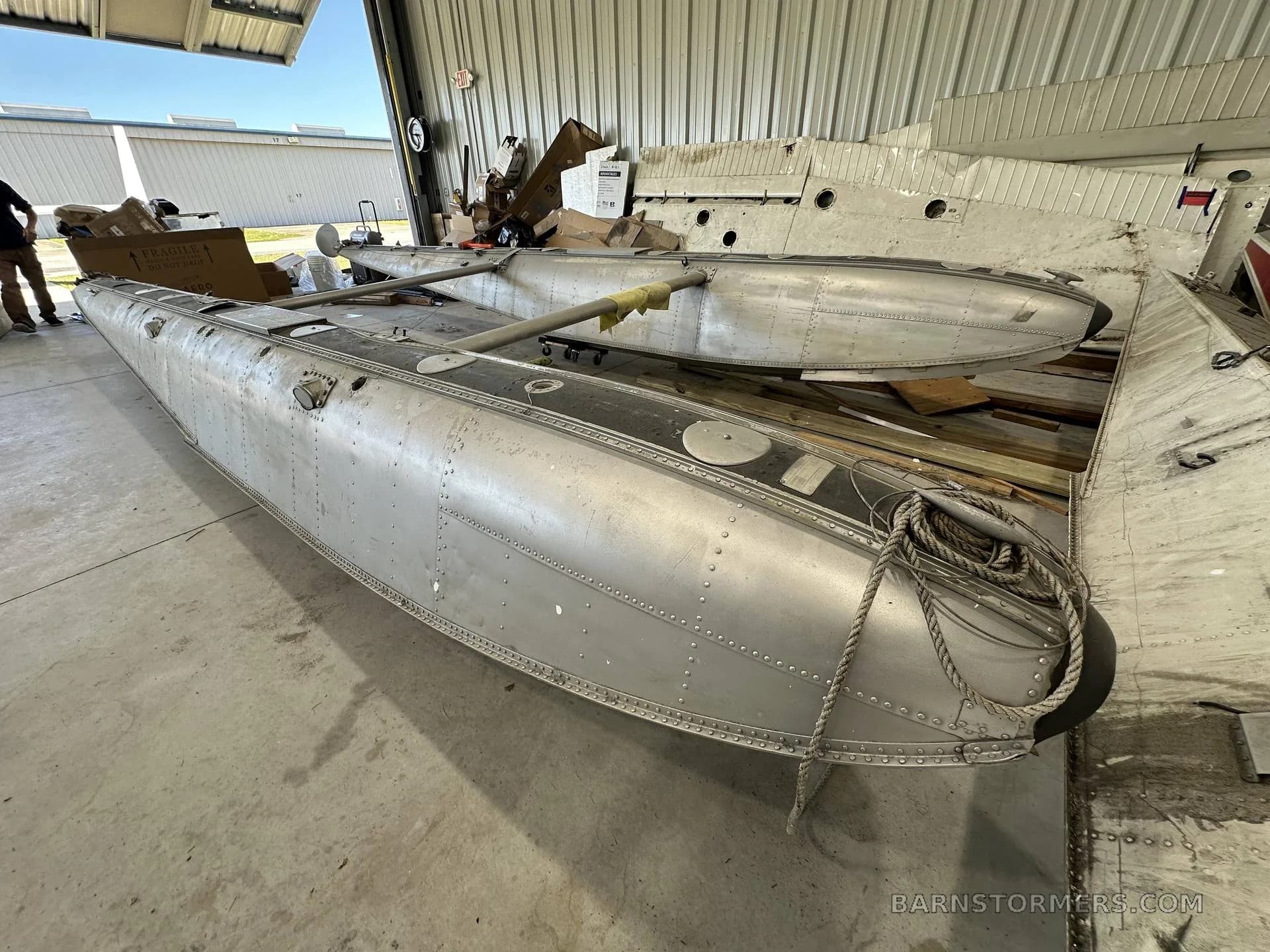 Two large aluminum aircraft floats are stored in a hangar, with one float partially disassembled.