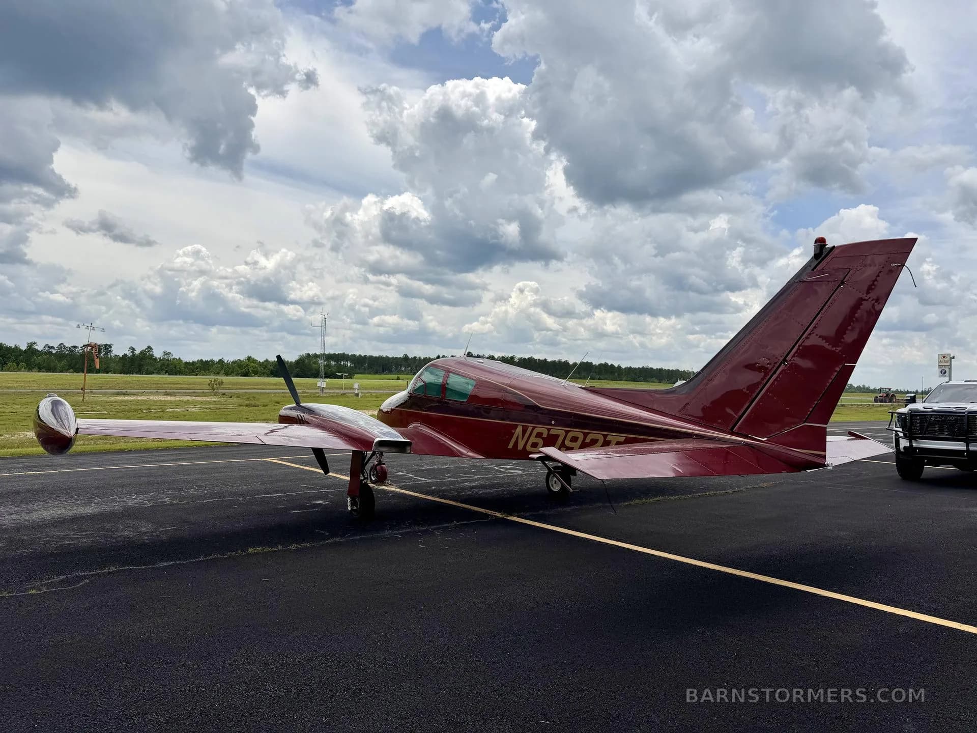 A rear-quarter view of a red twin-engine aircraft, likely a Beechcraft Baron, with registration N6792T, parked on an asphalt runway with a distant field.