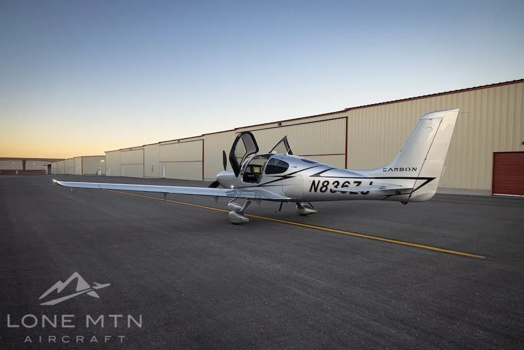 A silver Cirrus SR22T aircraft with registration N836ZJ is parked on an asphalt ramp with its doors open, viewed from the rear, with hangars in the background.