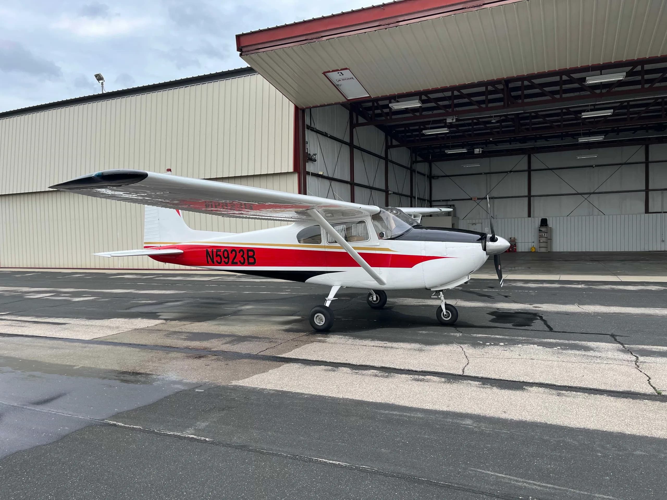 A side view of a white Cessna 182 aircraft with red and black stripes and registration N5923B, parked outside an open hangar door.