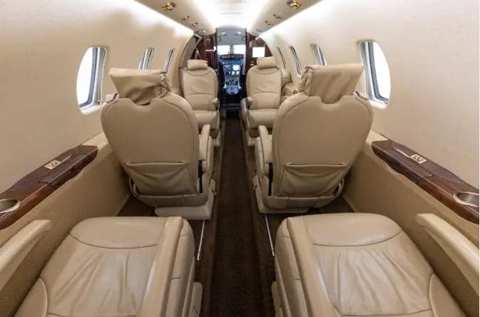 The spacious cabin interior of a business jet, with beige leather seats arranged in a club configuration, looking towards the cockpit.