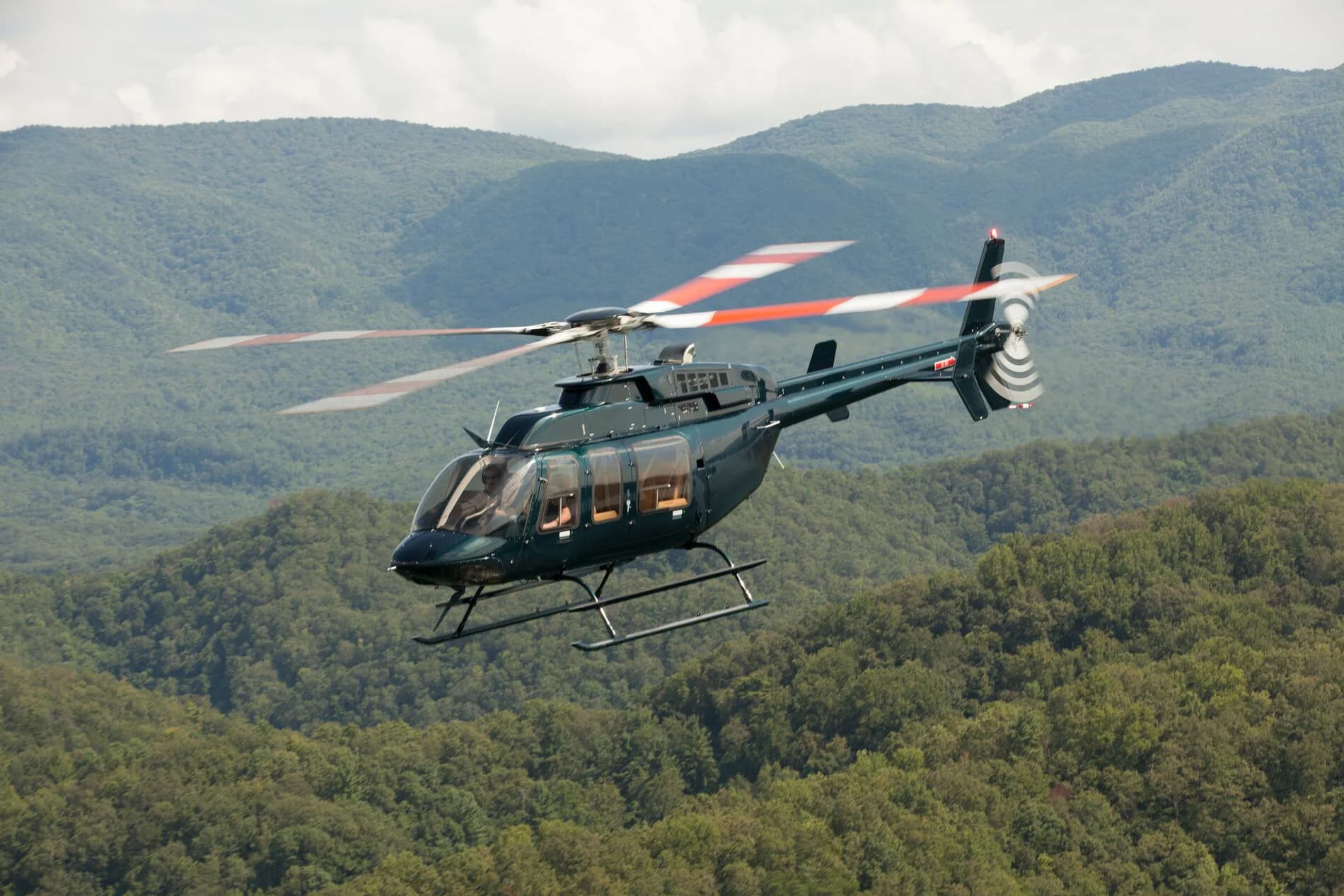 A dark green helicopter with red-striped main rotor blades flies over lush green mountains.