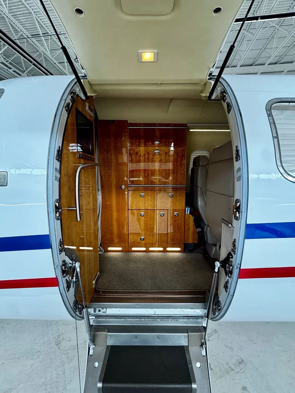 The open cabin door of the aircraft reveals a luxurious interior with polished wood cabinetry and a small galley area.